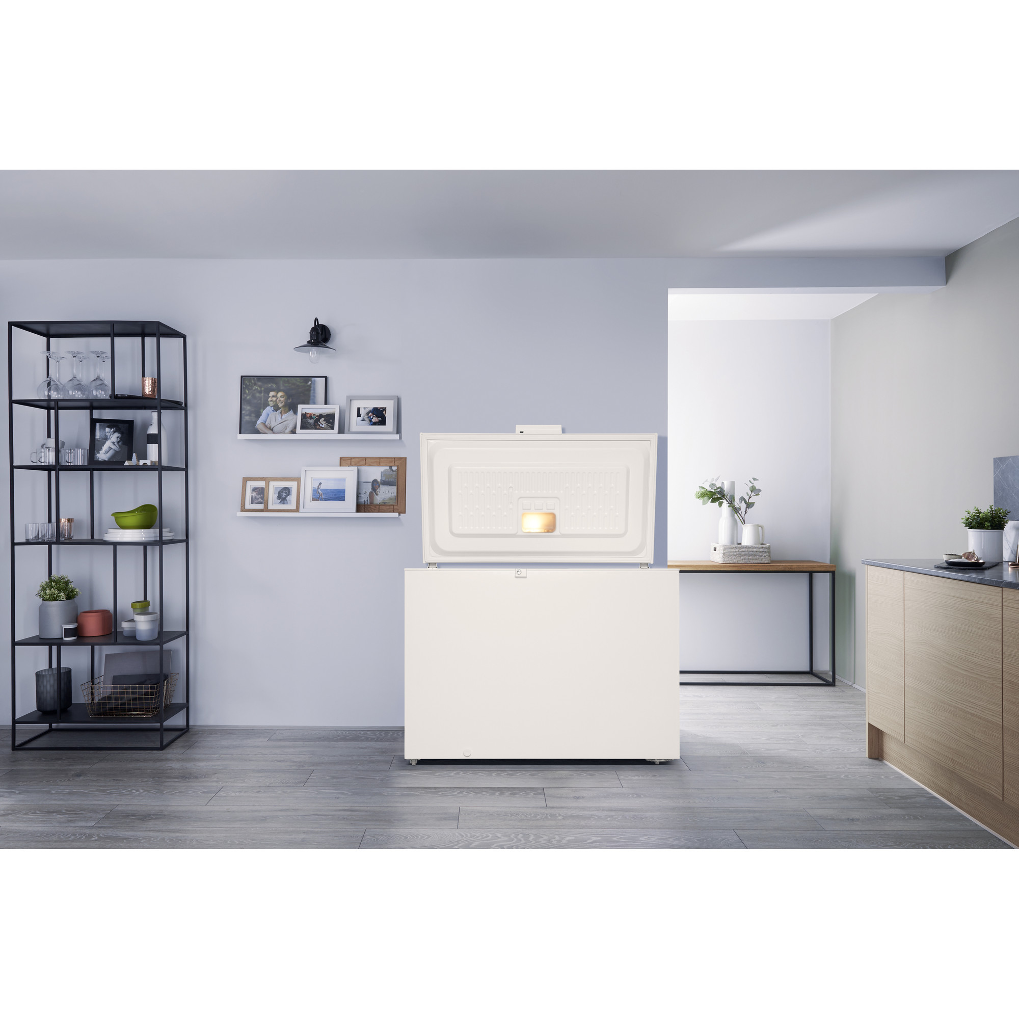 Ein weißer Gefrierschrank mit offener Tür. Ein Regal mit Gegenständen und ein Tisch mit Pflanzen im Hintergrund.