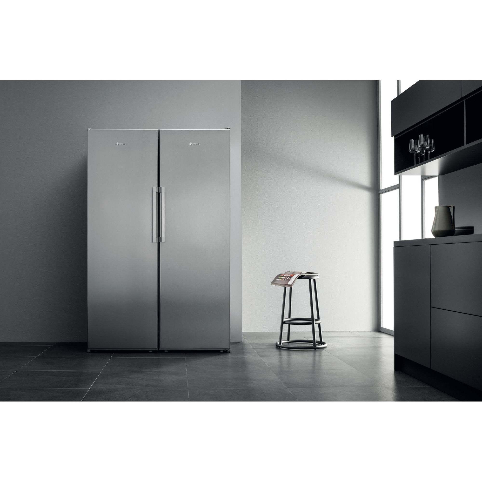 Ein Edelstahl-Kühlschrank mit nebeneinanderliegenden Türen in einem Raum mit Hocker und schwarzem Schrank.