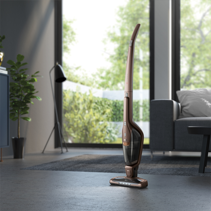 Ein bronzefarbener kabelloser Staubsauger steht in einem Wohnzimmer. Ein Fenster mit Grün im Hintergrund. Graues Sofa und Teppich.