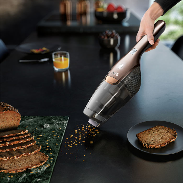 Handstaubsauger reinigt Krümel von einem schwarzen Tisch. Brot, Saft und Obst sind im Hintergrund.