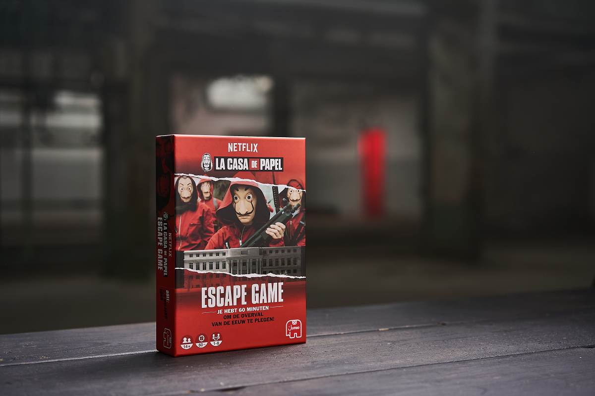 Een rode 'La Casa de Papel' Escape Game doos staat op een houten tafel. De achtergrond is wazig en toont een gebouw.