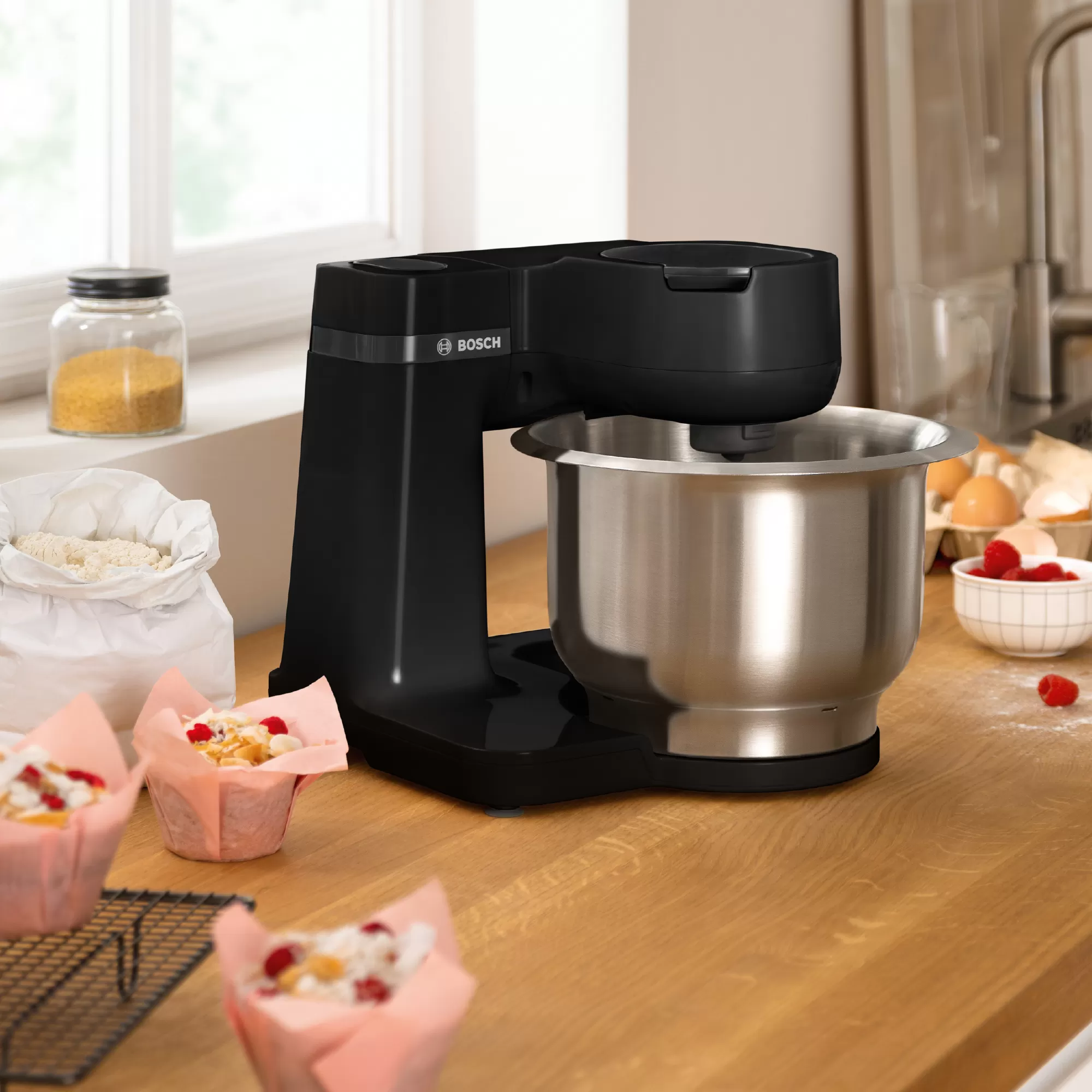 Robot pâtissier noir avec un bol en acier inoxydable et des muffins en arrière-plan.