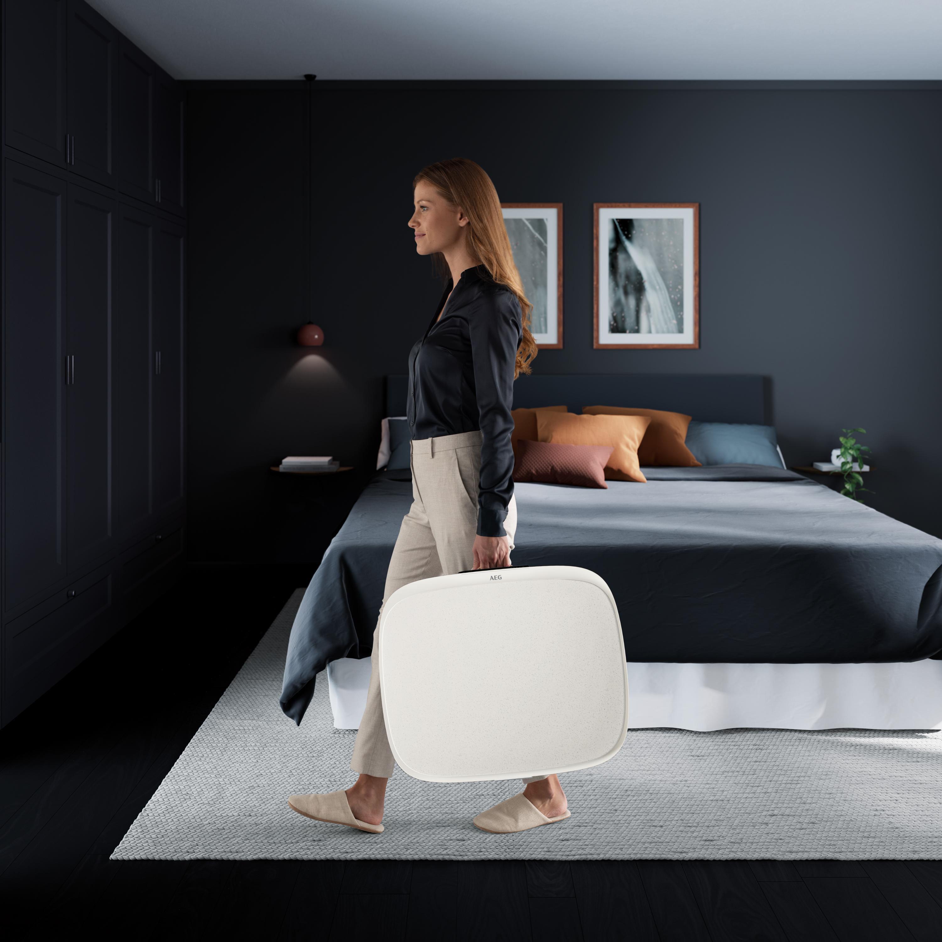 Eine Frau trägt einen weißen Koffer in einem Schlafzimmer. Dunkle Möbel, Bett im Hintergrund.