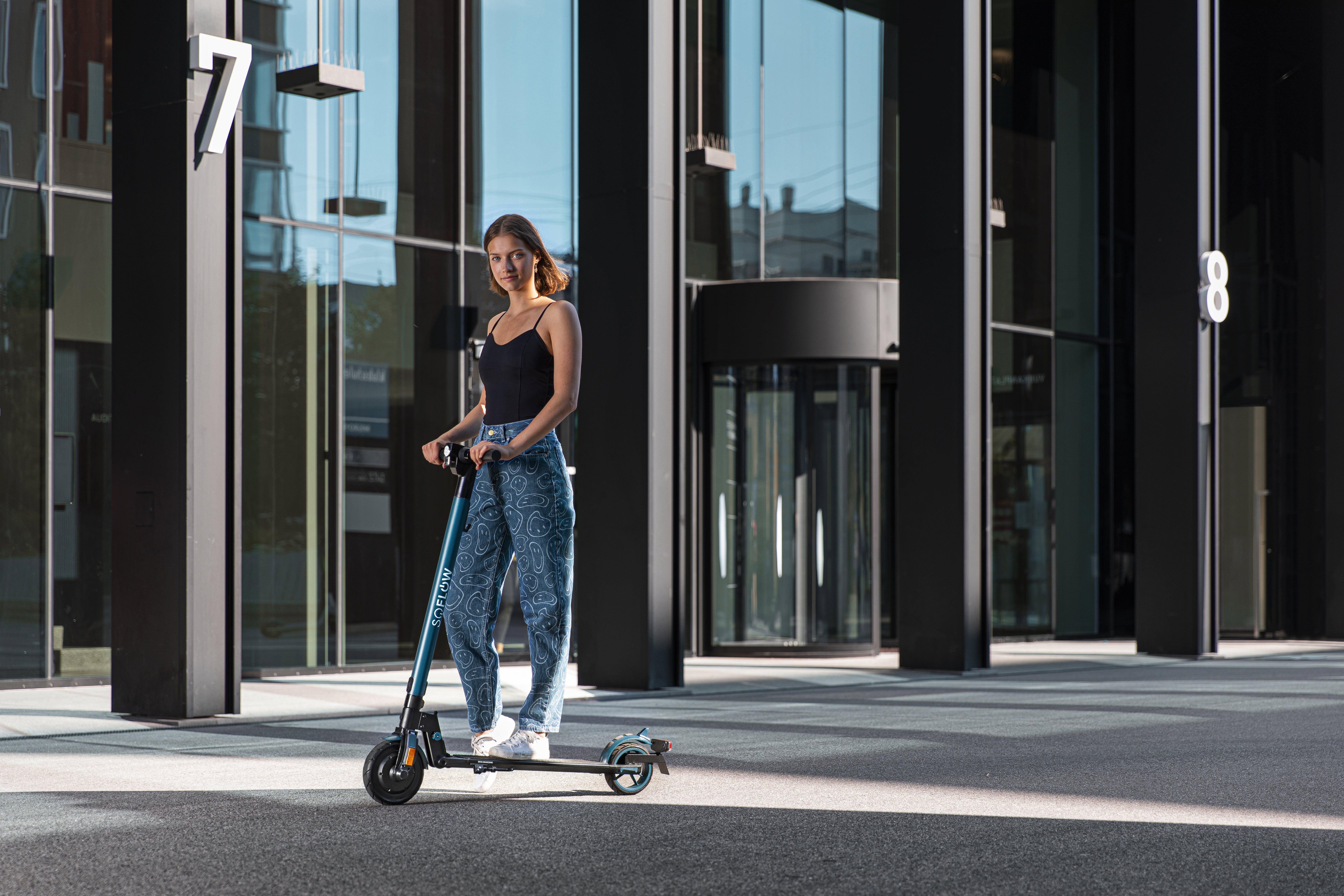 Frau steht vor einem Gebäude auf einem Roller. Sie trägt Jeans und ein schwarzes Oberteil.