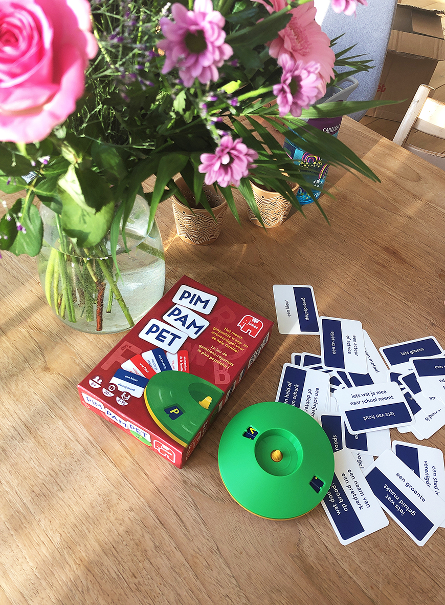Een tafel met een speldoos, kaarten en een groene spinner. Een vaas met roze bloemen en potplanten zijn ook aanwezig.