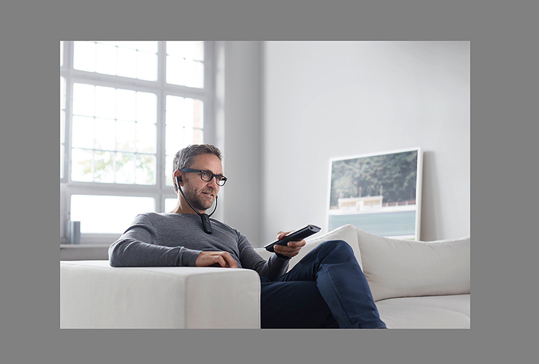 Man in glasses, holding a remote, sitting on a white couch.