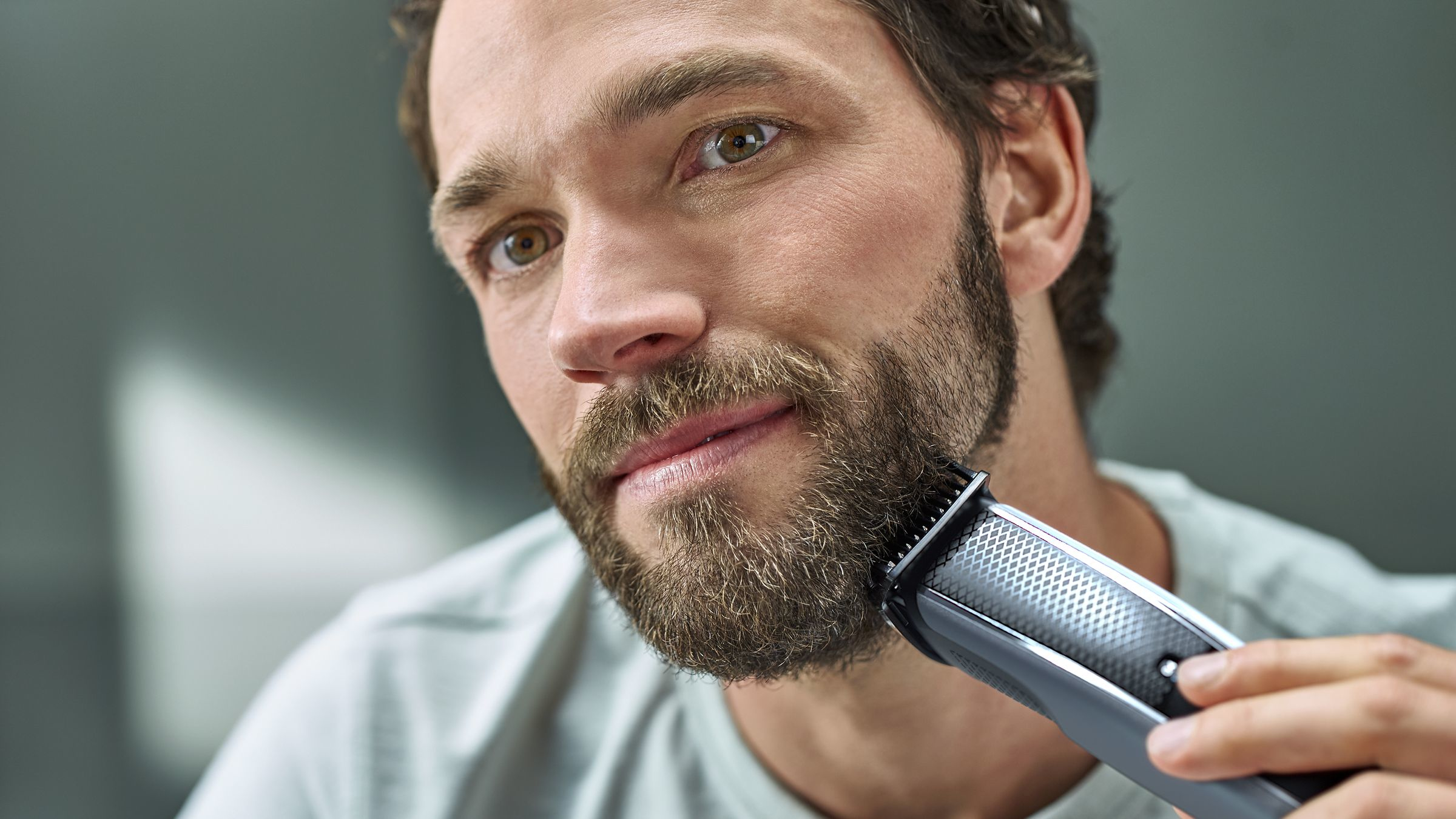 Een man scheert zijn baard met een trimmer. Hij heeft bruin haar, ogen en een volle baard. De achtergrond is grijs.