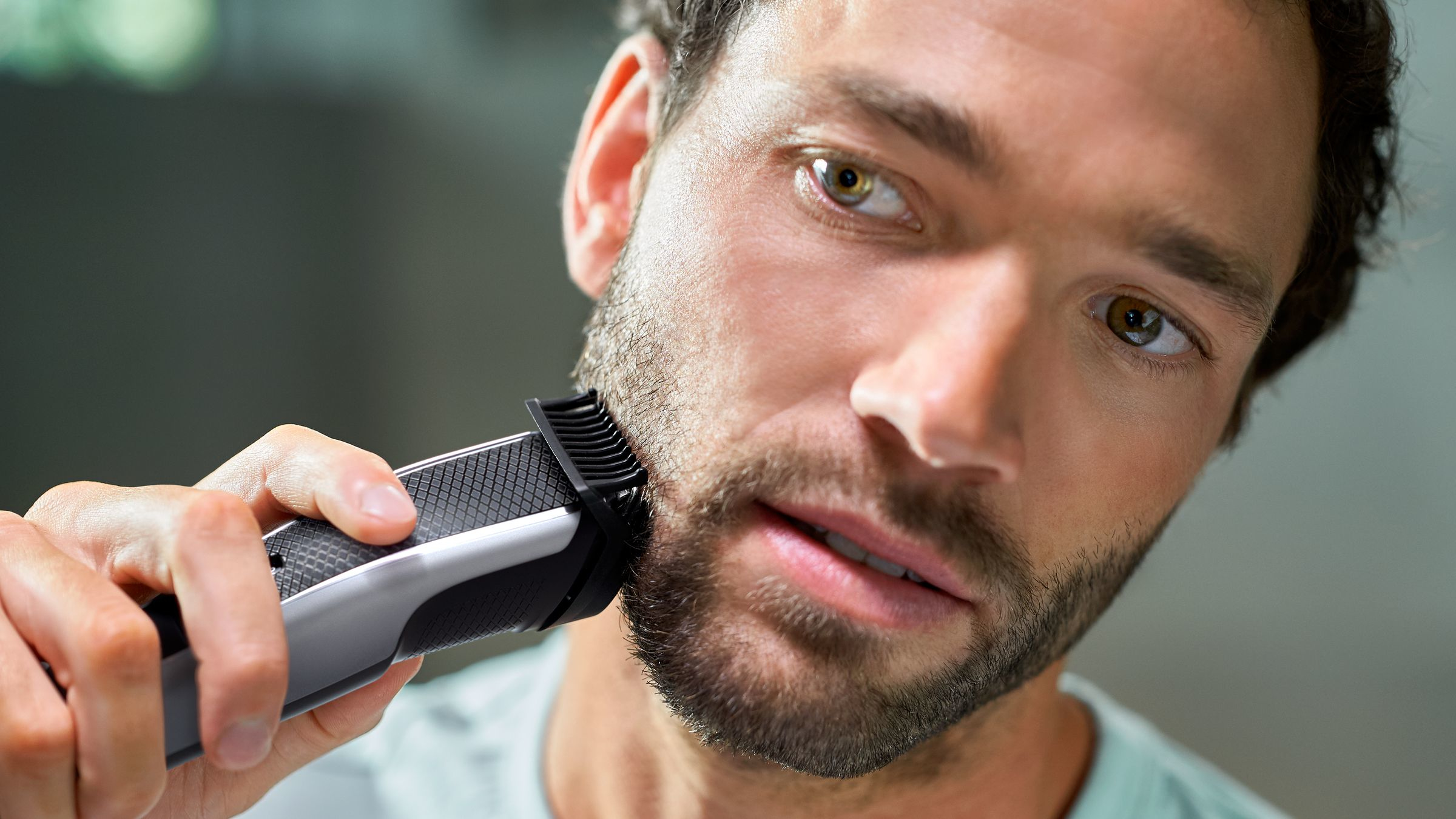 Een man scheert zijn baard met een elektrisch scheerapparaat. De man heeft bruin haar en ogen. Het scheerapparaat is zwart en zilver.