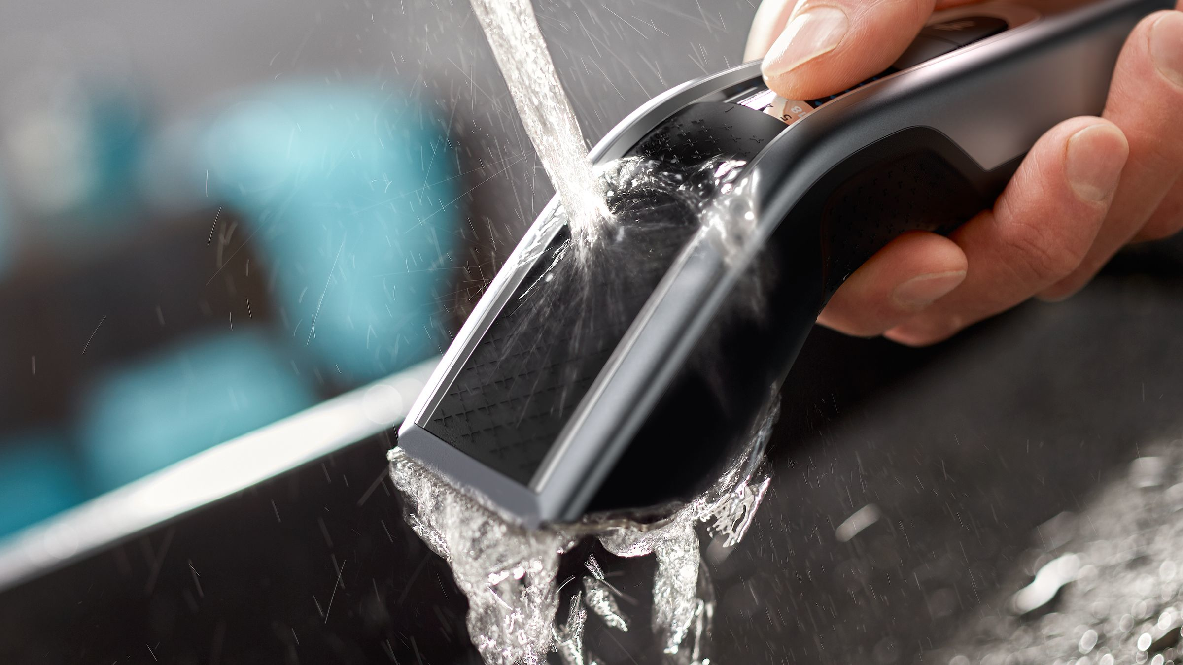 Water is pouring over a black and gray Philips hair clipper, held by a hand. Water is splashing.