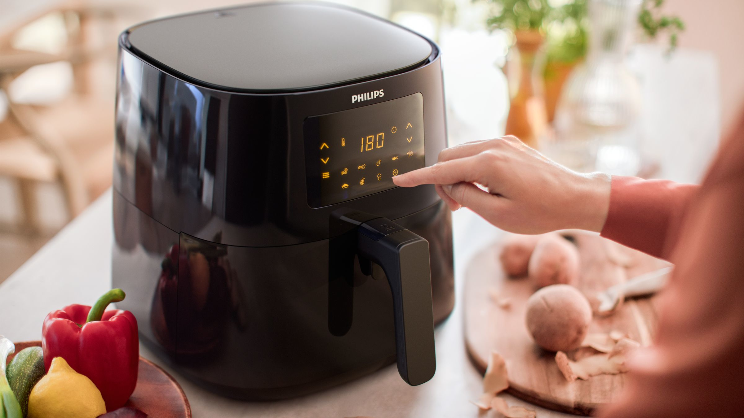 Zwarte airfryer met digitaal display en hand die het aanraakt, groenten op de achtergrond.