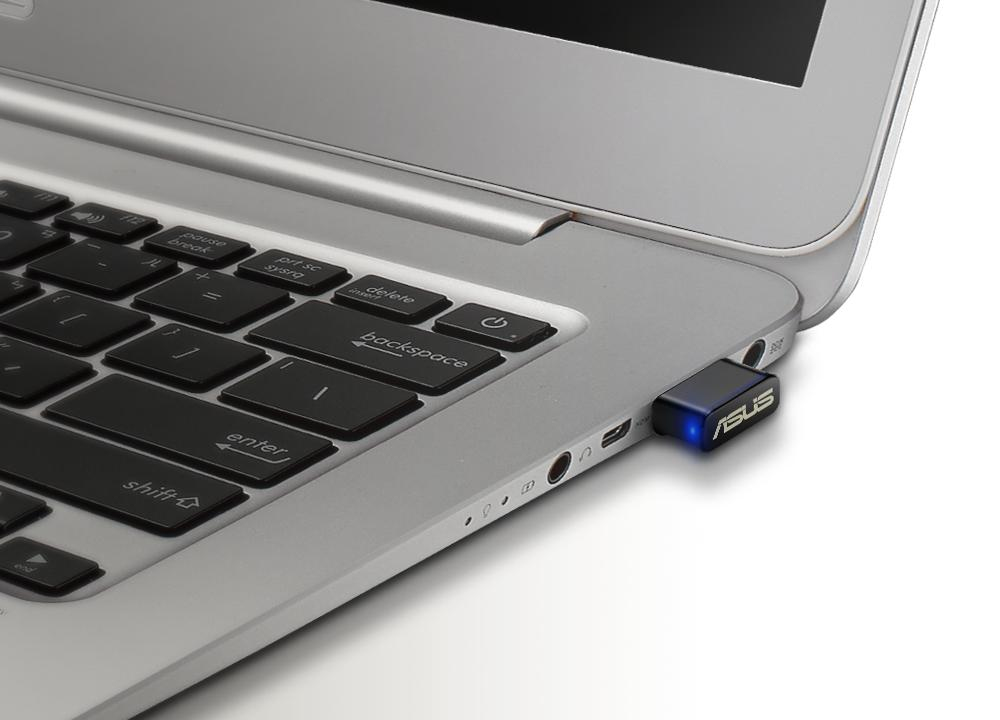 Un portátil plateado con un dongle USB negro insertado, logotipo ASUS iluminado, sobre superficie blanca.