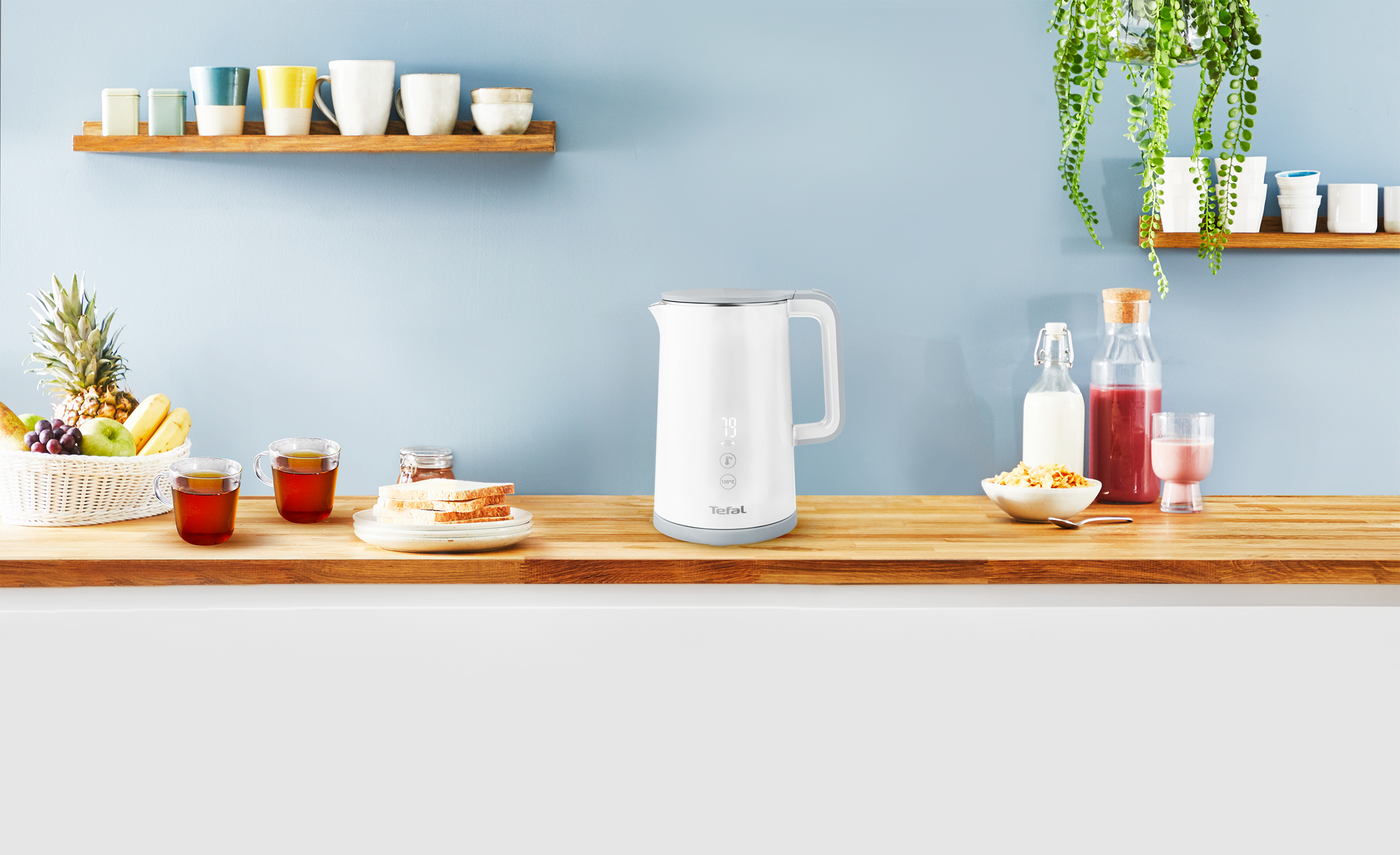 Weißer Wasserkocher auf einer Holz-Arbeitsplatte. Küche mit Essen und Dekorationen im Hintergrund.