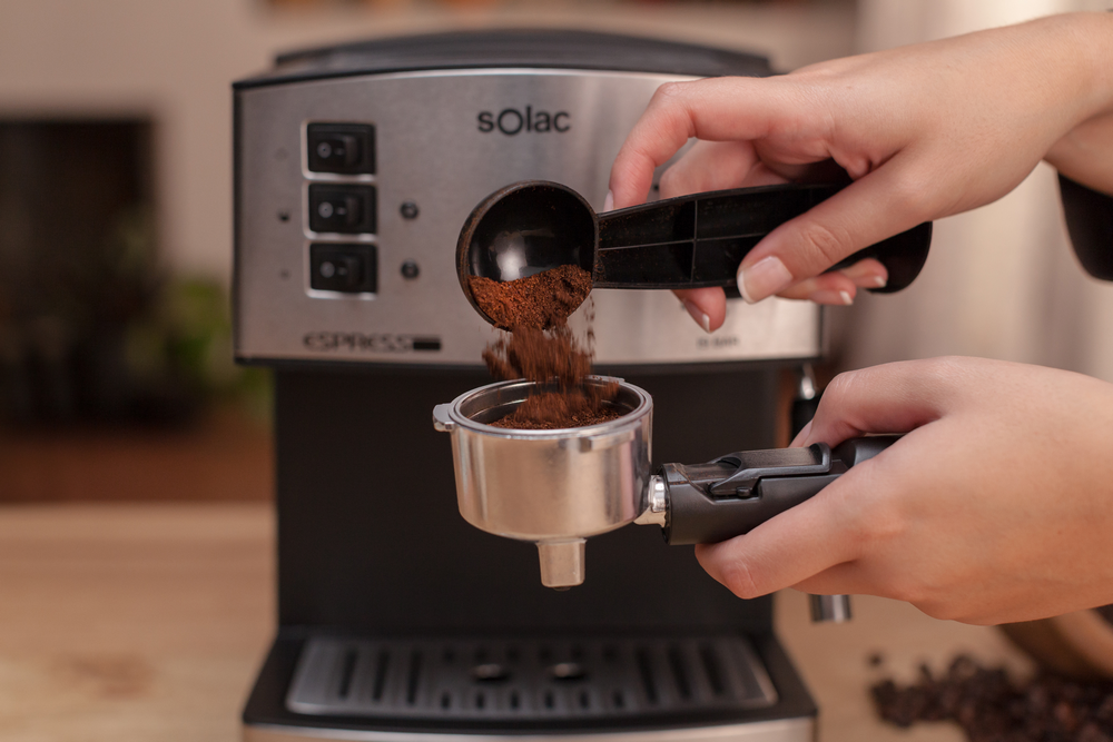 Manos vertiendo café molido en un portafiltro sostenido en una máquina de espresso.