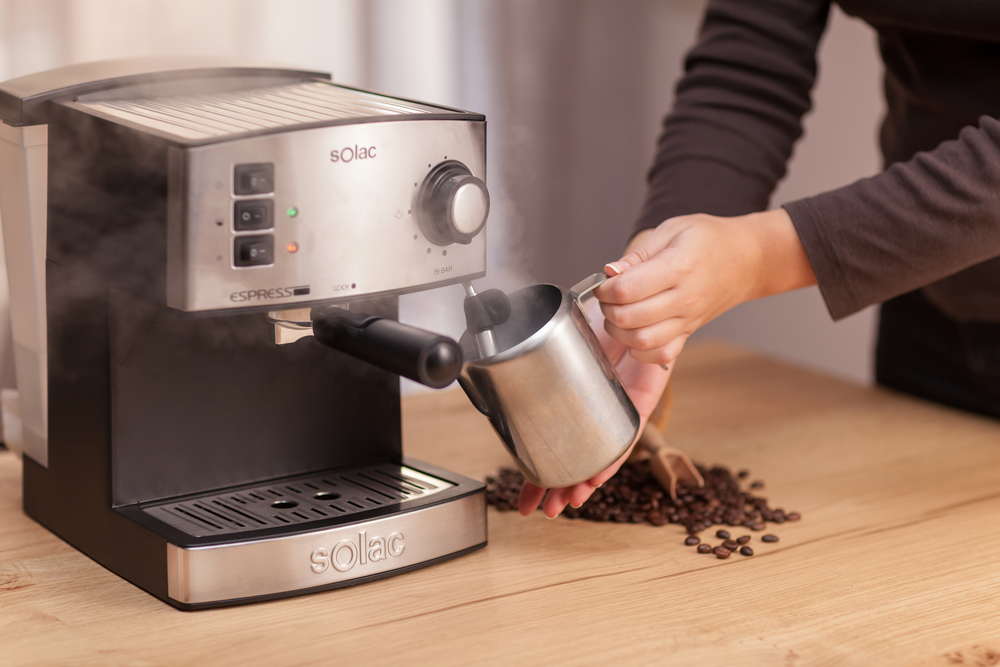 Máquina de espresso echando vapor en jarra de leche de acero inoxidable, granos de café en mesa.
