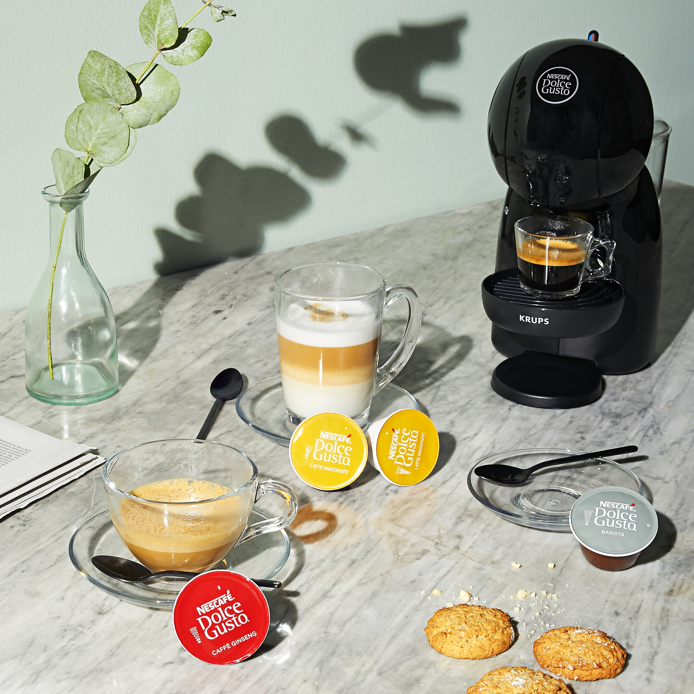 Une machine à café avec des tasses et des capsules de café. Un verre avec du café en couches et des biscuits sur une table en marbre.