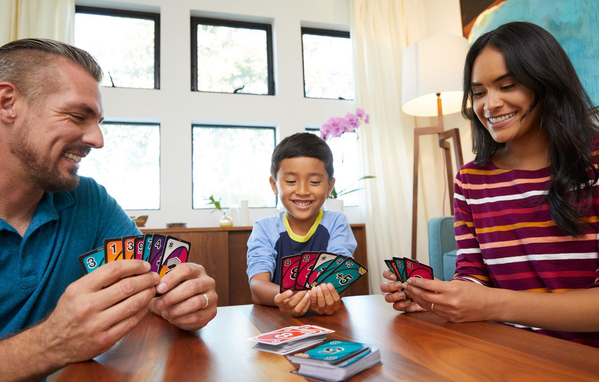Familie spielt Uno-Kartenspiel. Drei Personen mit Karten um einen Tisch im Wohnzimmer.