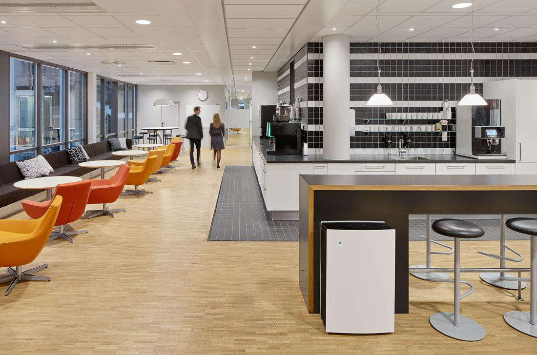 Ein Büro mit einem langen Tisch, Stühlen und Personen, die gehen. Ein großer Luftreiniger ist vorhanden.
