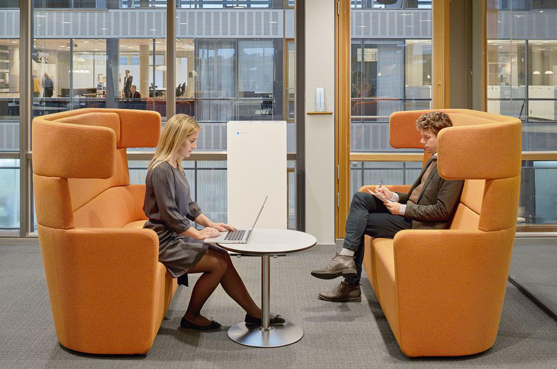 Zwei Personen in einem modernen Büro, die in orangefarbenen Kabinen mit einem weißen Luftreiniger sitzen.