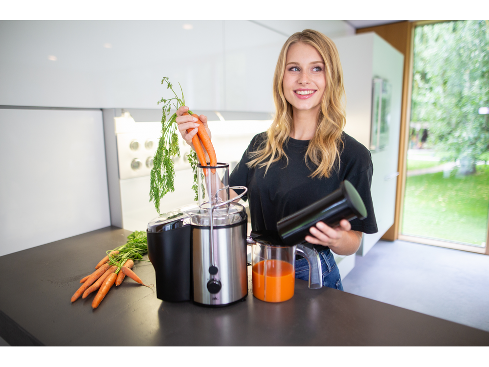 Mujer agregando zanahorias a una exprimidora. Zanahorias y jugo en primer plano. Interior de cocina en el fondo.