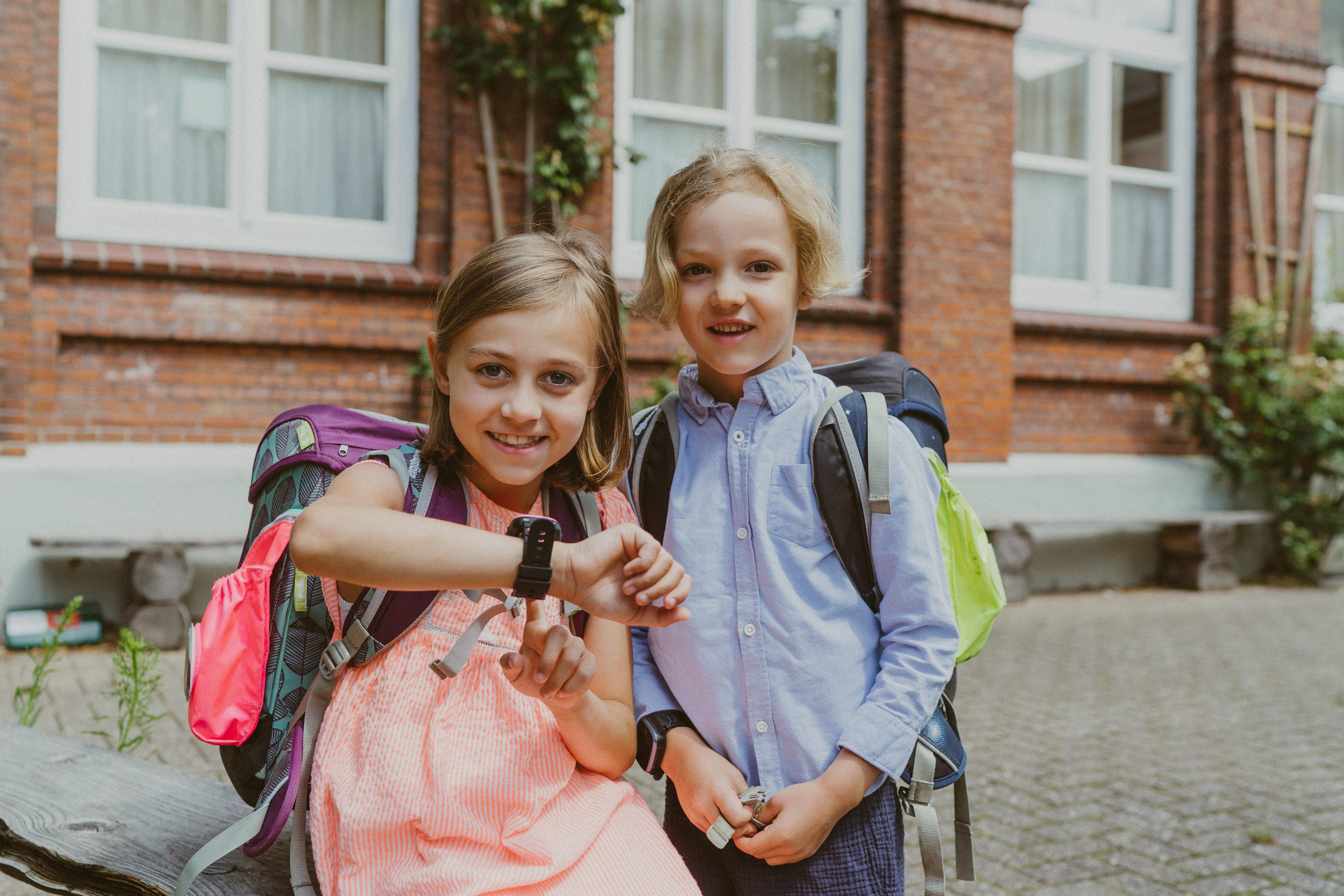 Zwei Kinder mit Rucksäcken, eines schaut auf eine Smartwatch, vor einem Backsteingebäude.