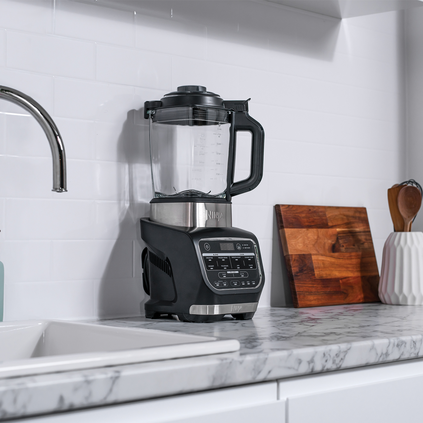 Ein Ninja-Mixer steht auf einer Marmorarbeitsplatte. Ein Schneidebrett und Utensilien befinden sich rechts. Im Vordergrund sind ein Waschbecken und ein Wasserhahn zu sehen.