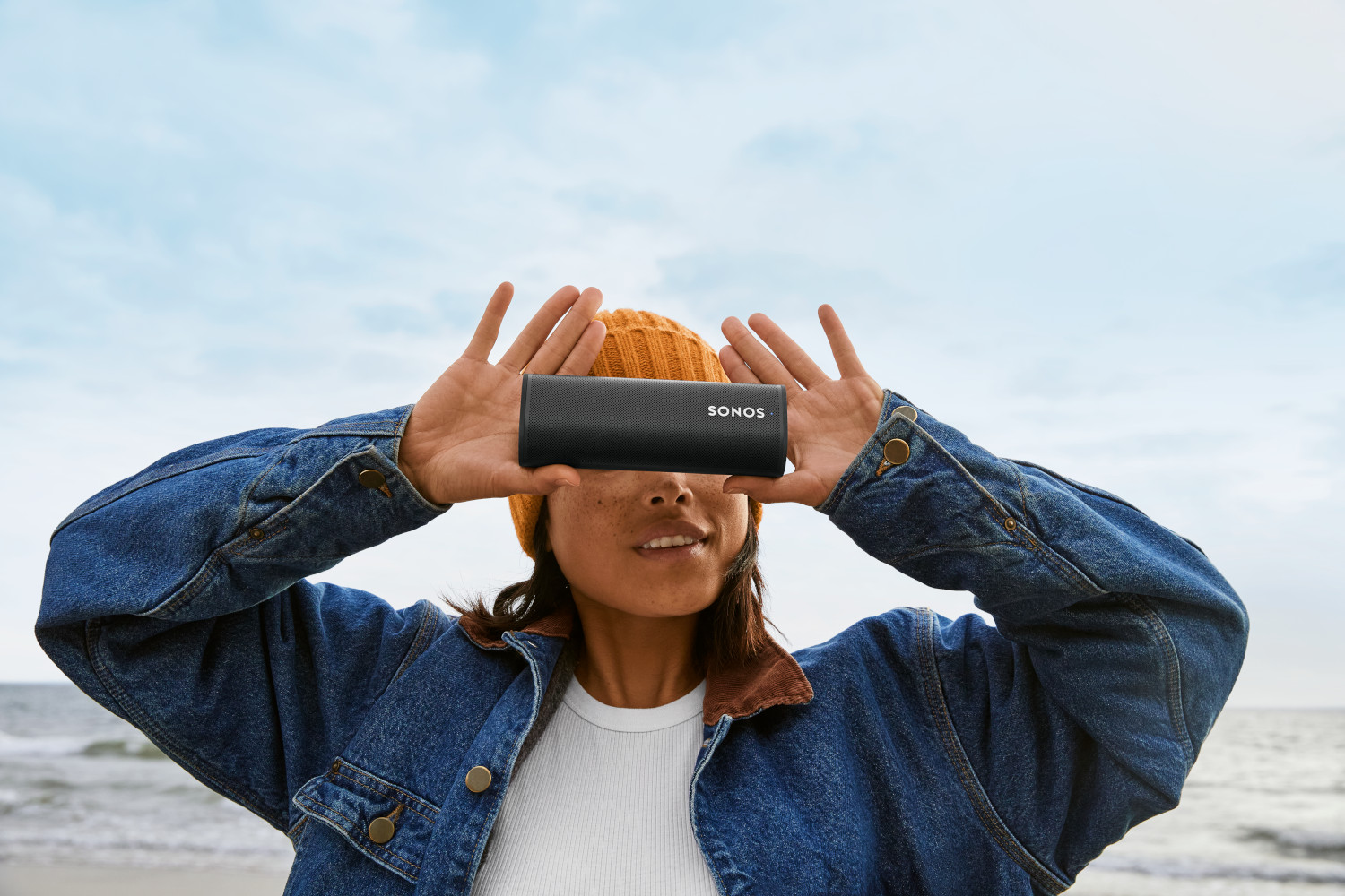 Een vrouw die een zwarte Sonos-luidspreker voor haar ogen houdt, op het strand.