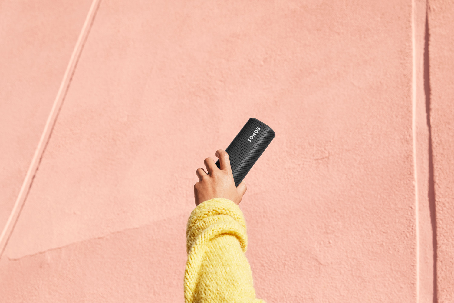 Een hand die een zwarte Sonos-luidspreker vasthoudt tegen een roze muur.