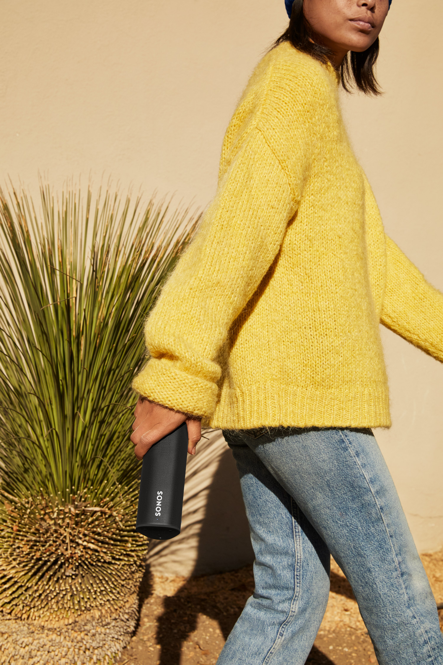 Een persoon in gele trui en jeans, houdt een zwarte Sonos-luidspreker vast, buiten lopend.