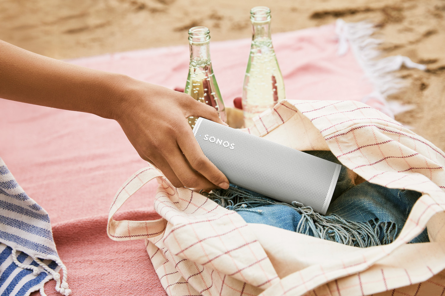 Une main plongeant dans un sac pour sortir un haut-parleur Sonos blanc sur une plage.