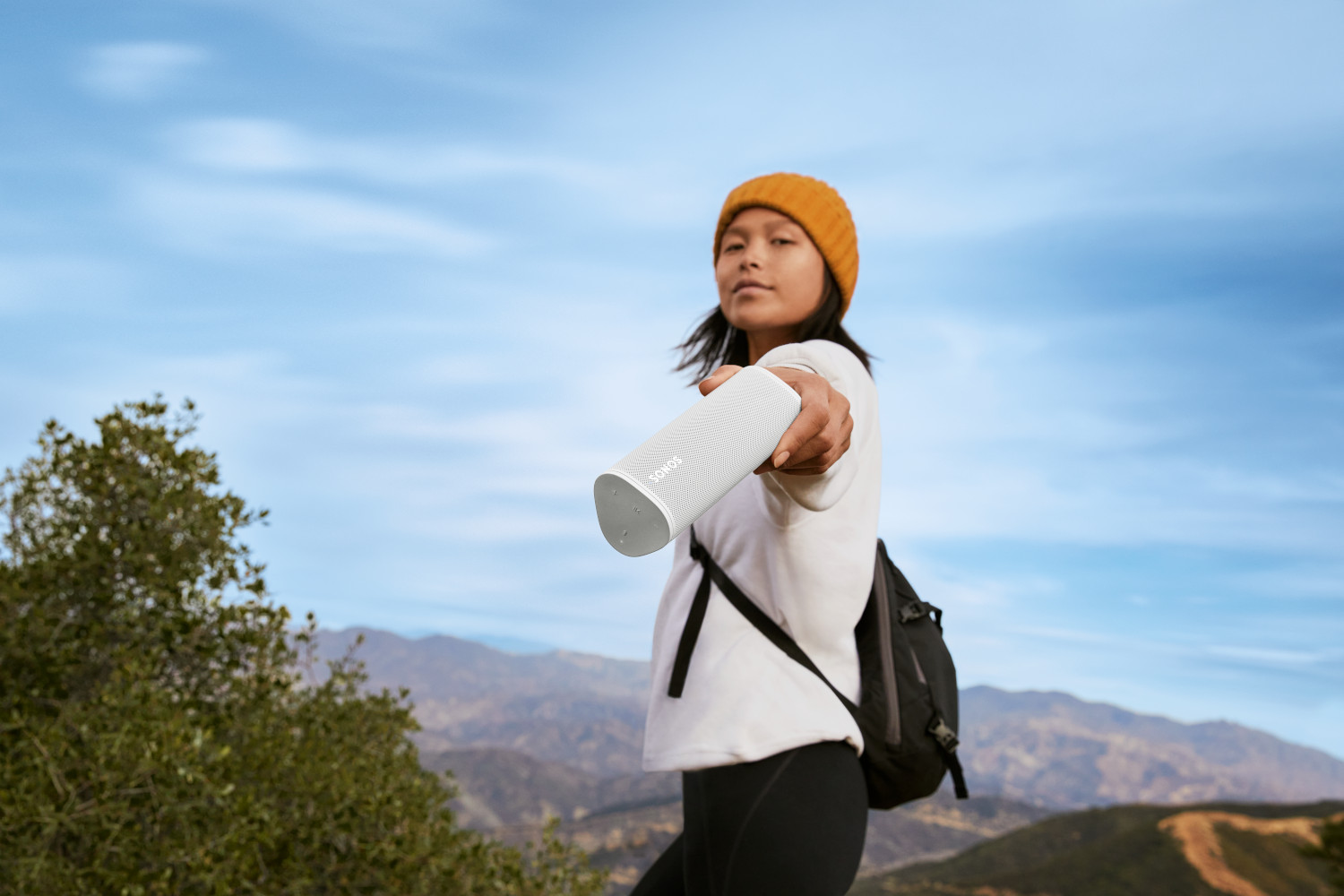 Une femme avec un chapeau tenant un haut-parleur blanc, avec un paysage en arrière-plan.