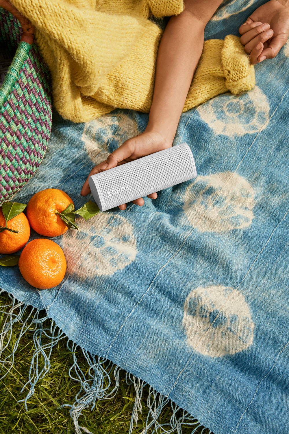 Une personne tenant un haut-parleur Sonos blanc à côté d'oranges et d'une couverture bleue.