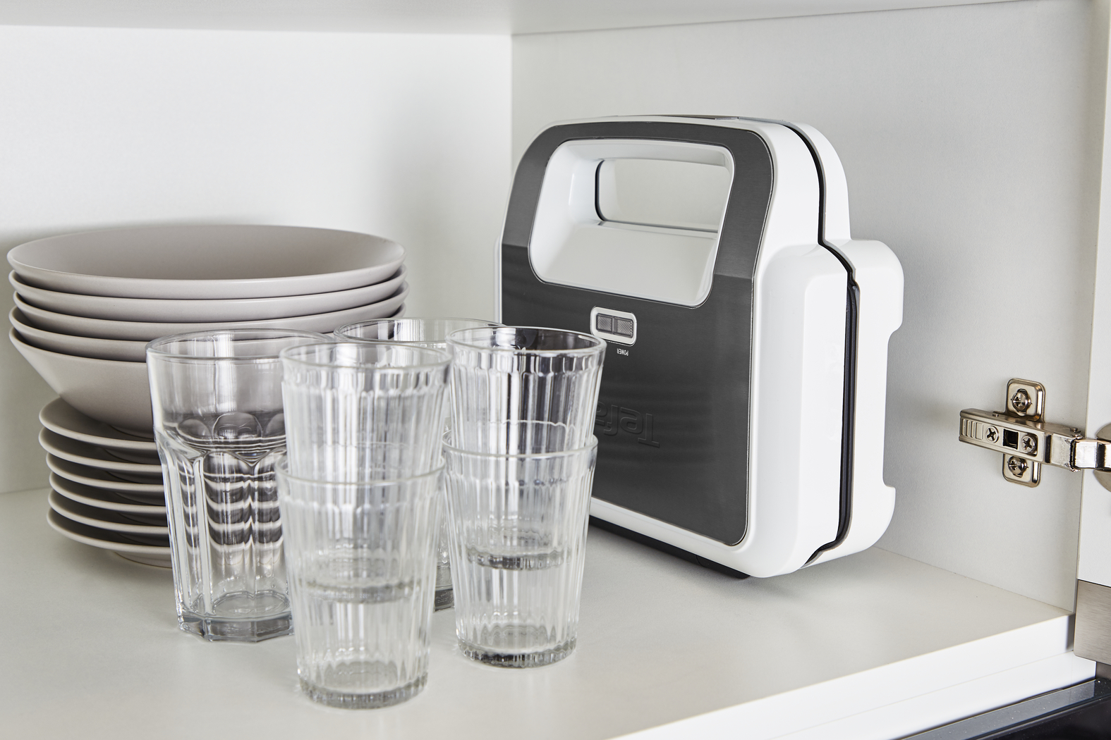 White, white-and-gray waffle maker and several glasses and plates in a cabinet.