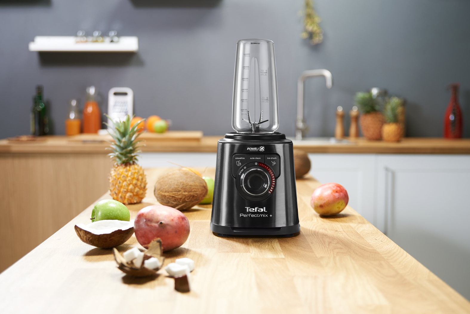Een zwarte blender met fruit op een houten tafel. Een ananas, kokosnoot en mango zijn zichtbaar.