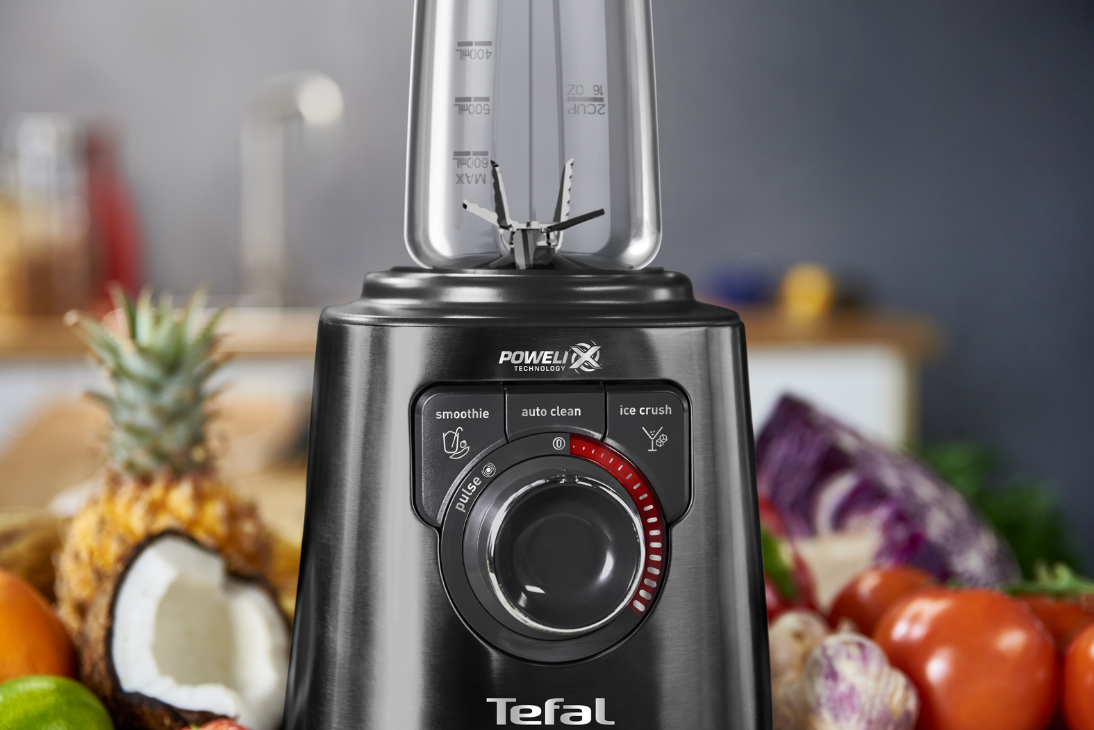 Zwarte en zilveren Tefal blender met fruit op de achtergrond. Heeft een bedieningsknop.