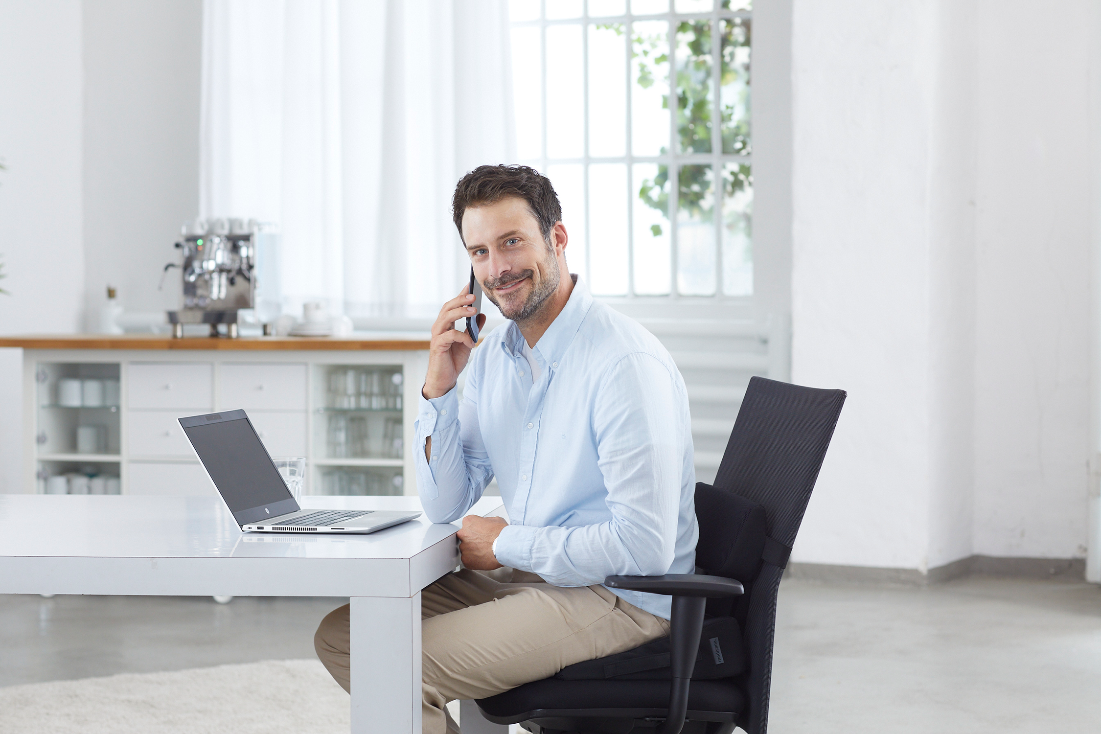 Een man zit aan een tafel, gebruikt een laptop, belt.