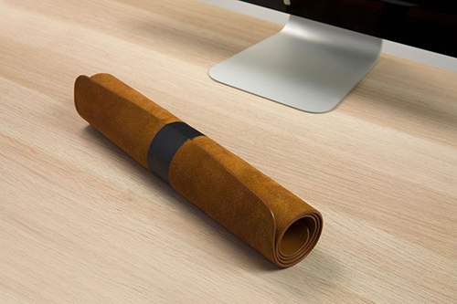 Een opgerolde bruine leren bureaumat met een zwarte band ligt op een houten bureau, in de buurt van een computermonitor.