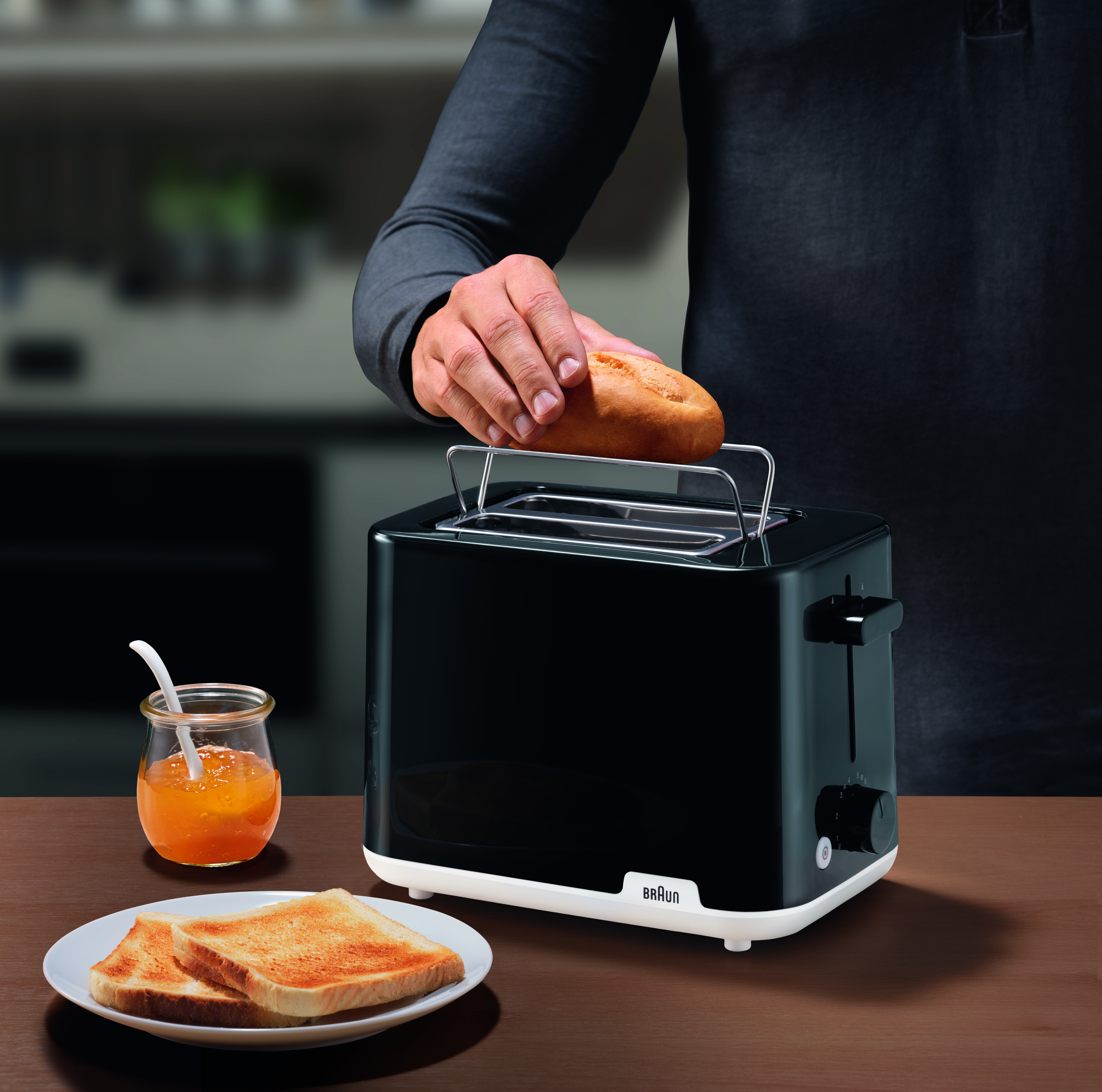 Eine Person legt Brot in einen schwarzen Braun-Toaster, mit Toast und Marmelade.