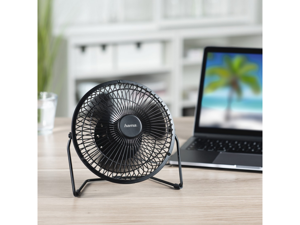 Un ventilador de escritorio negro sobre un escritorio de madera, con un portátil y un vaso de agua en el fondo.
