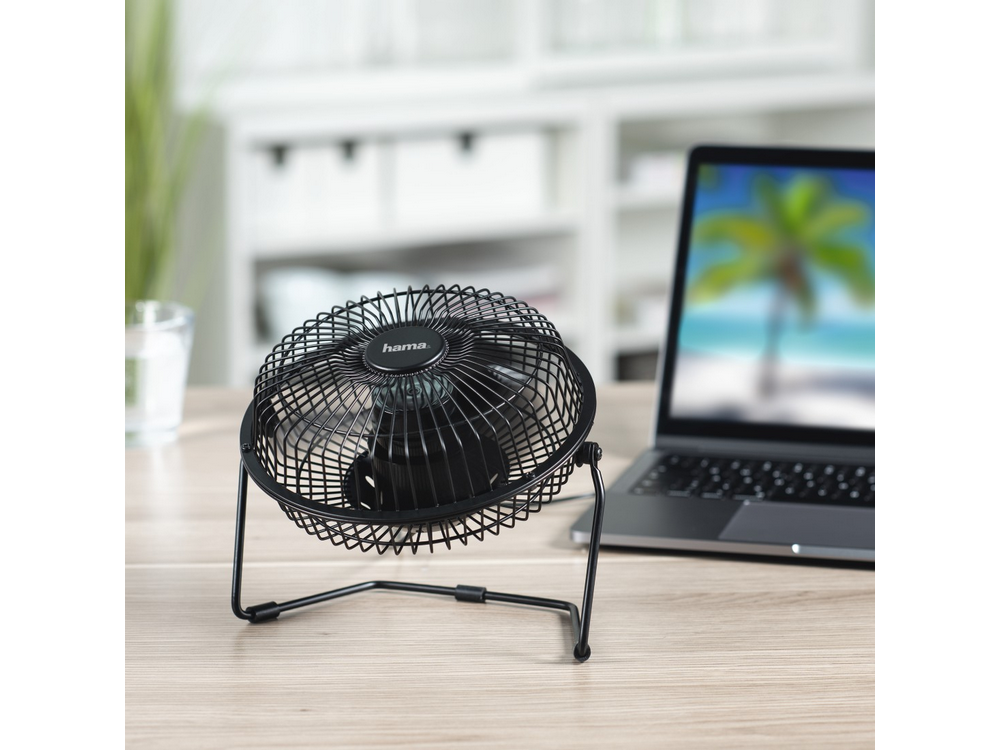 Ventilador de escritorio negro en un escritorio con laptop. El ventilador es negro con jaula protectora.