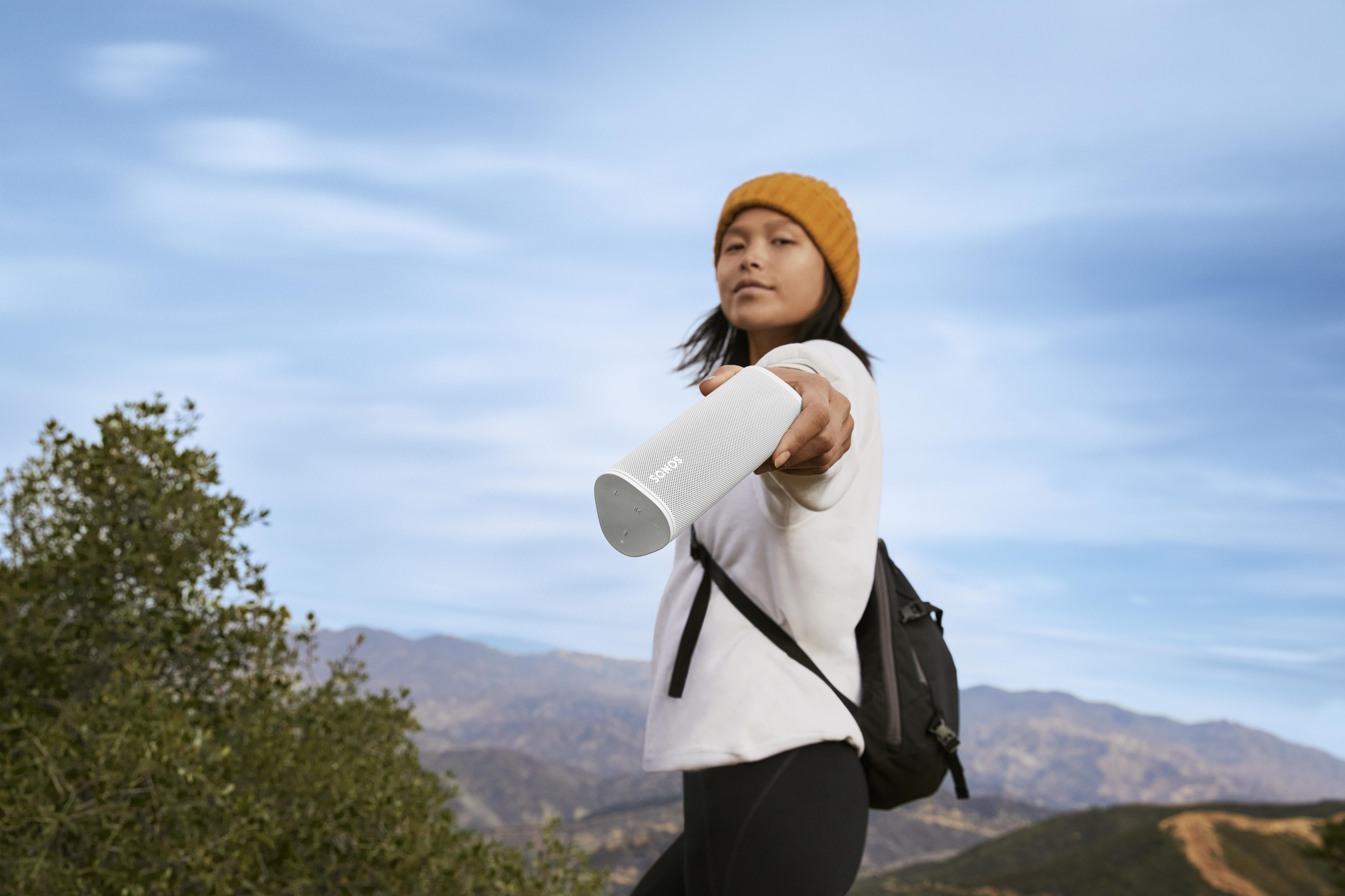 Frau hält einen weißen Sonos-Lautsprecher. Sie trägt einen Hut und einen Rucksack und posiert im Freien.