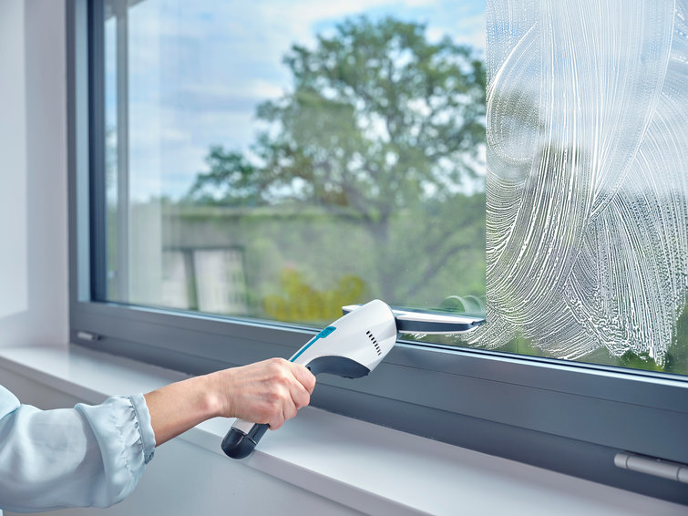 Eine Person putzt ein Fenster mit einem weißen und teal Fensterreiniger. Draußen sind Bäume und blauer Himmel sichtbar.