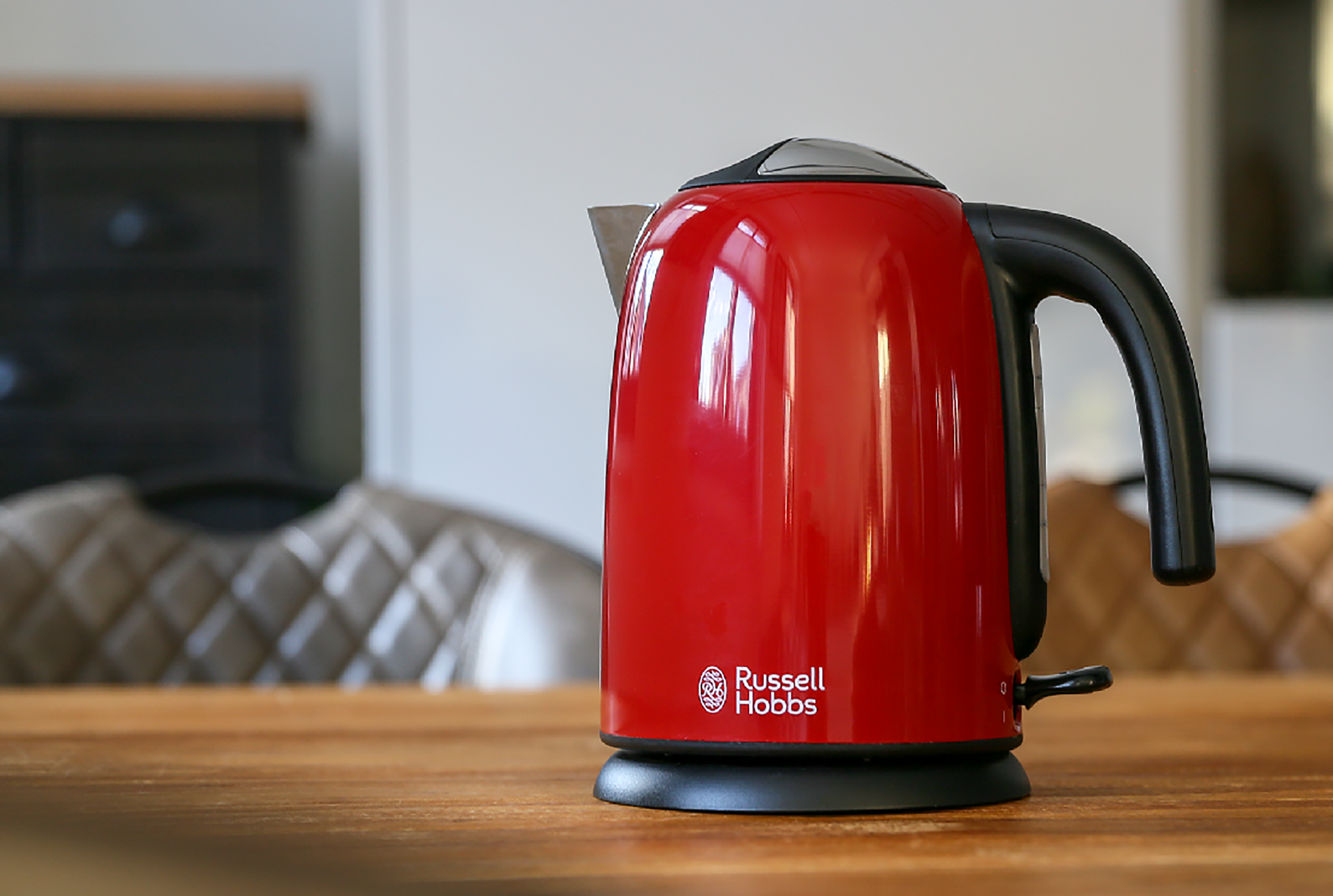 A red Russell Hobbs electric kettle sits on a wooden table, with a blurred background of a room.