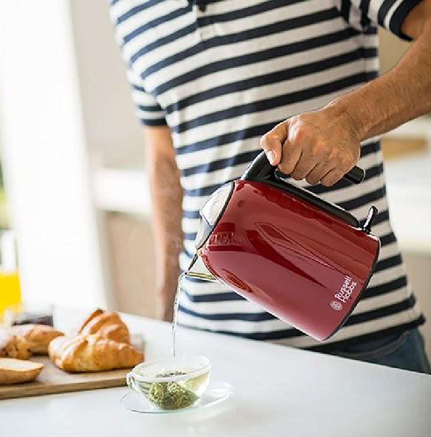 A person pouring tea from a red kettle into a teacup with pastries next to it.