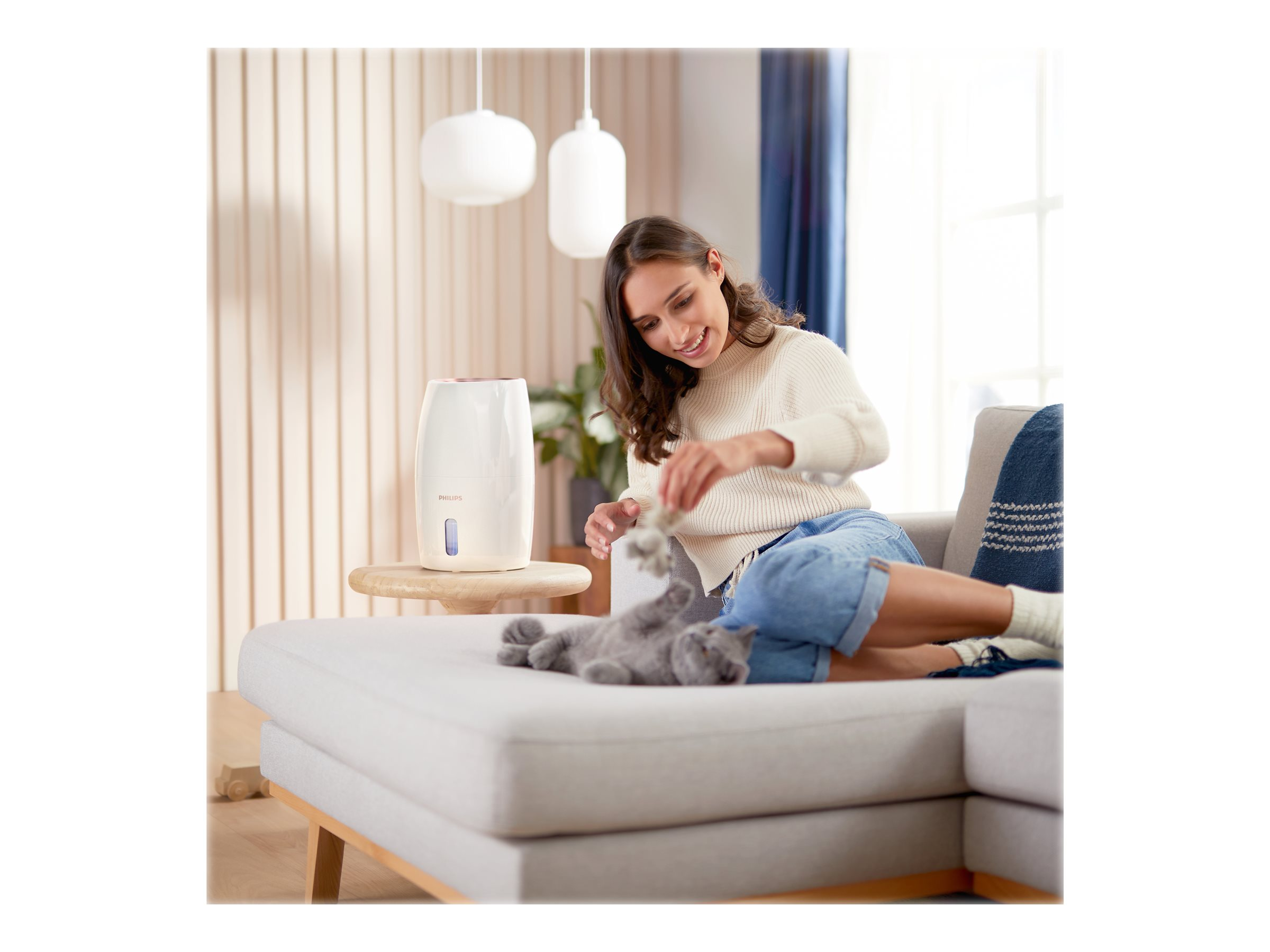Eine Frau sitzt auf einem Sofa und spielt mit einer grauen Katze. Ein Luftbefeuchter steht daneben.