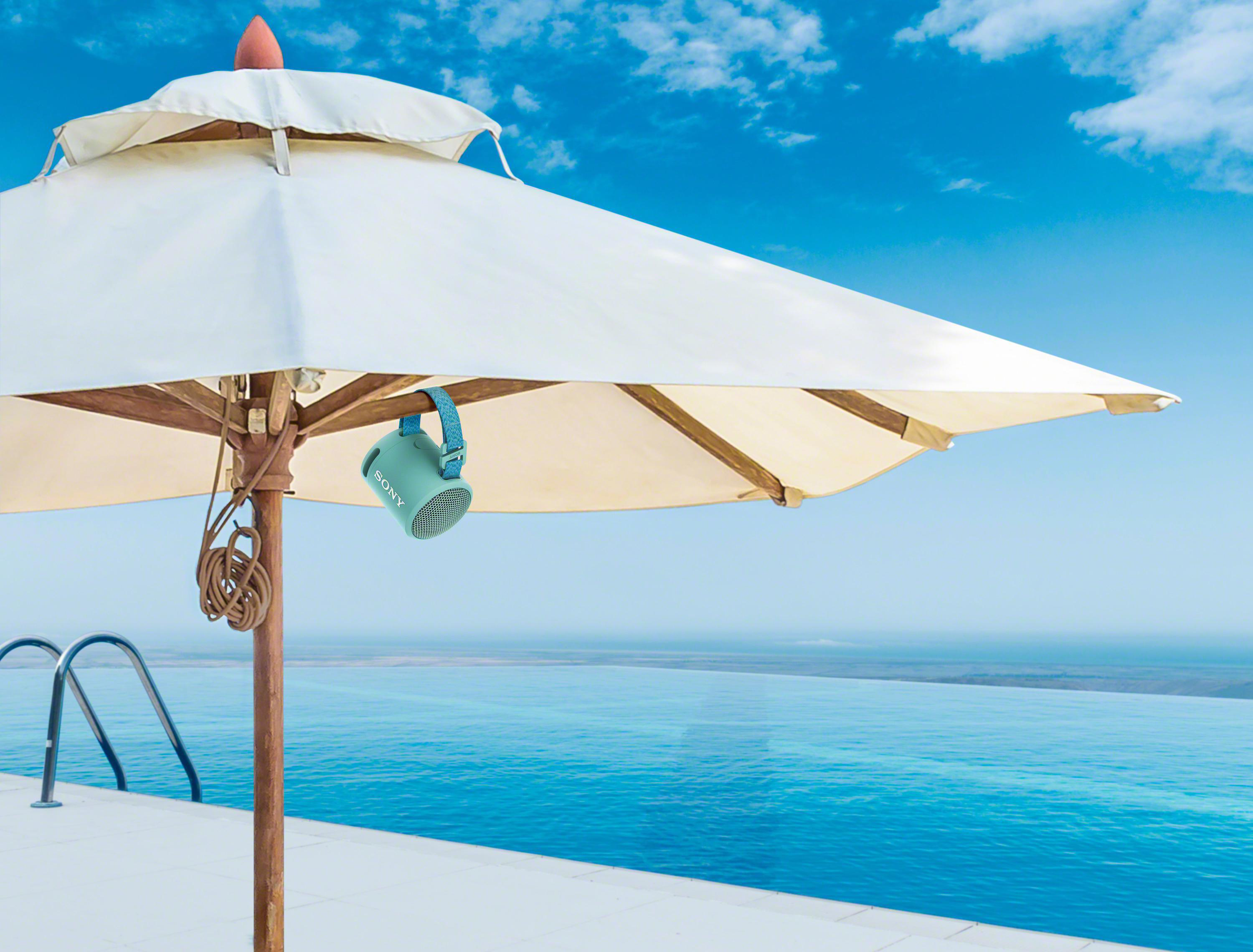 Ein offener Schirm mit einem hängenden türkisfarbenen Lautsprecher in der Nähe eines Infinity-Pools unter blauem Himmel.