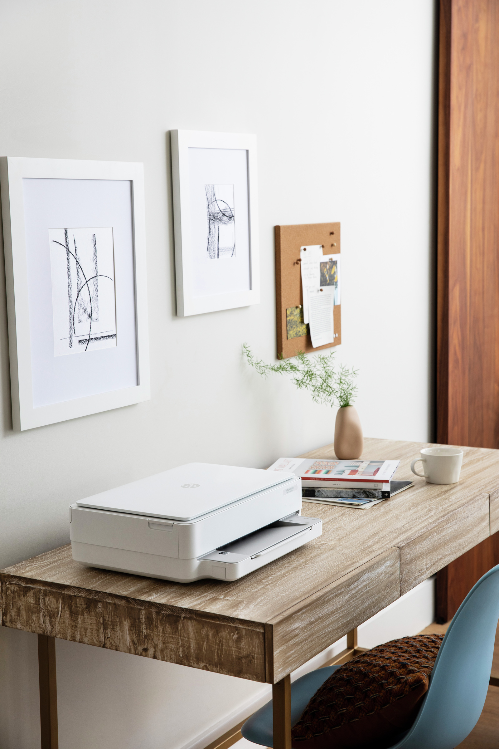 Una impresora blanca sobre un escritorio de madera con imágenes y un tablón de anuncios en la pared detrás.
