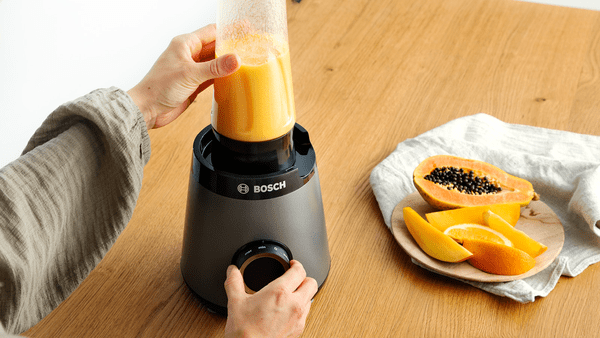 Een persoon mixt een smoothie met een blender. Papaya en sinaasappelschijfjes ernaast.