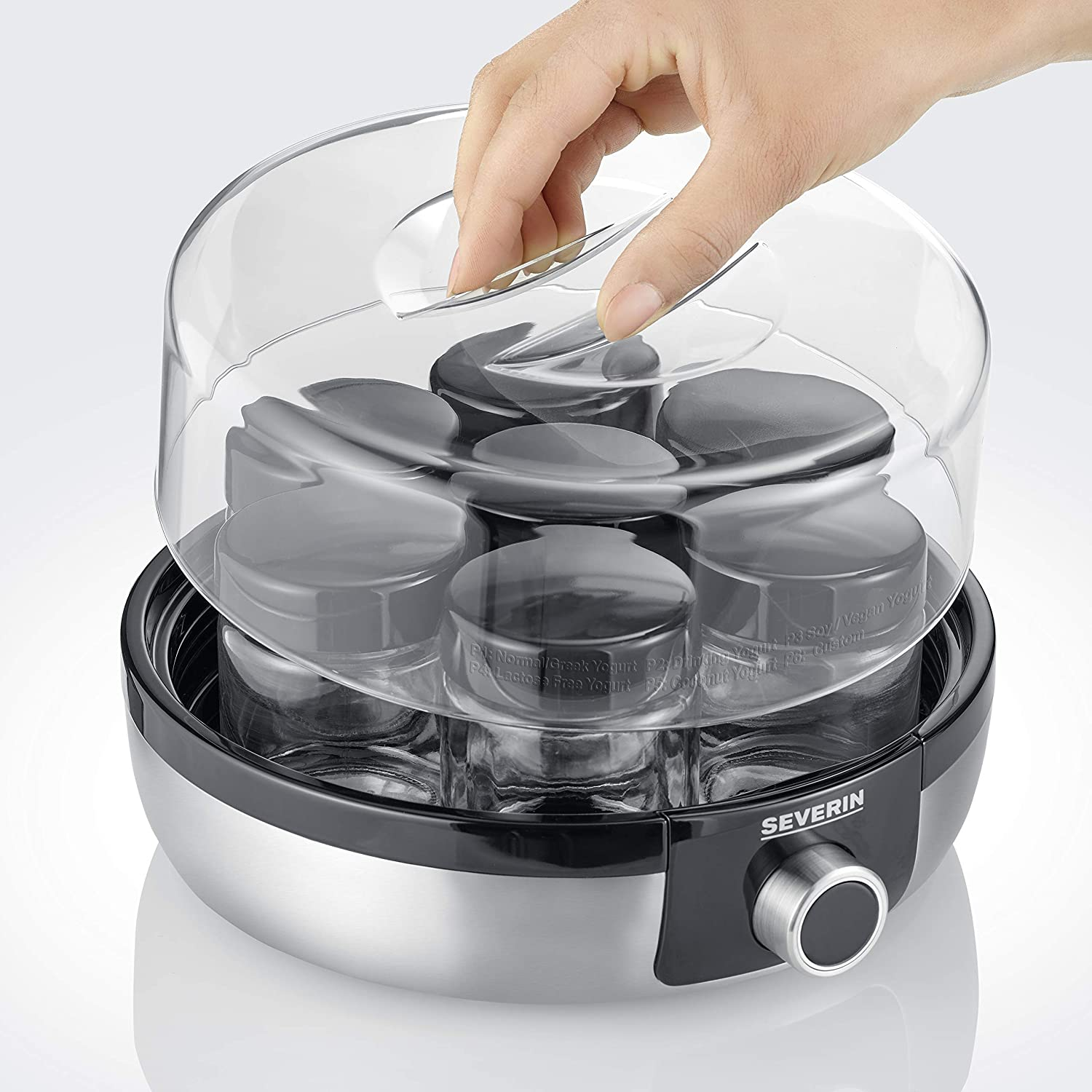 Een hand die het transparante deksel opent van een yoghurtmaker met glazen potten.