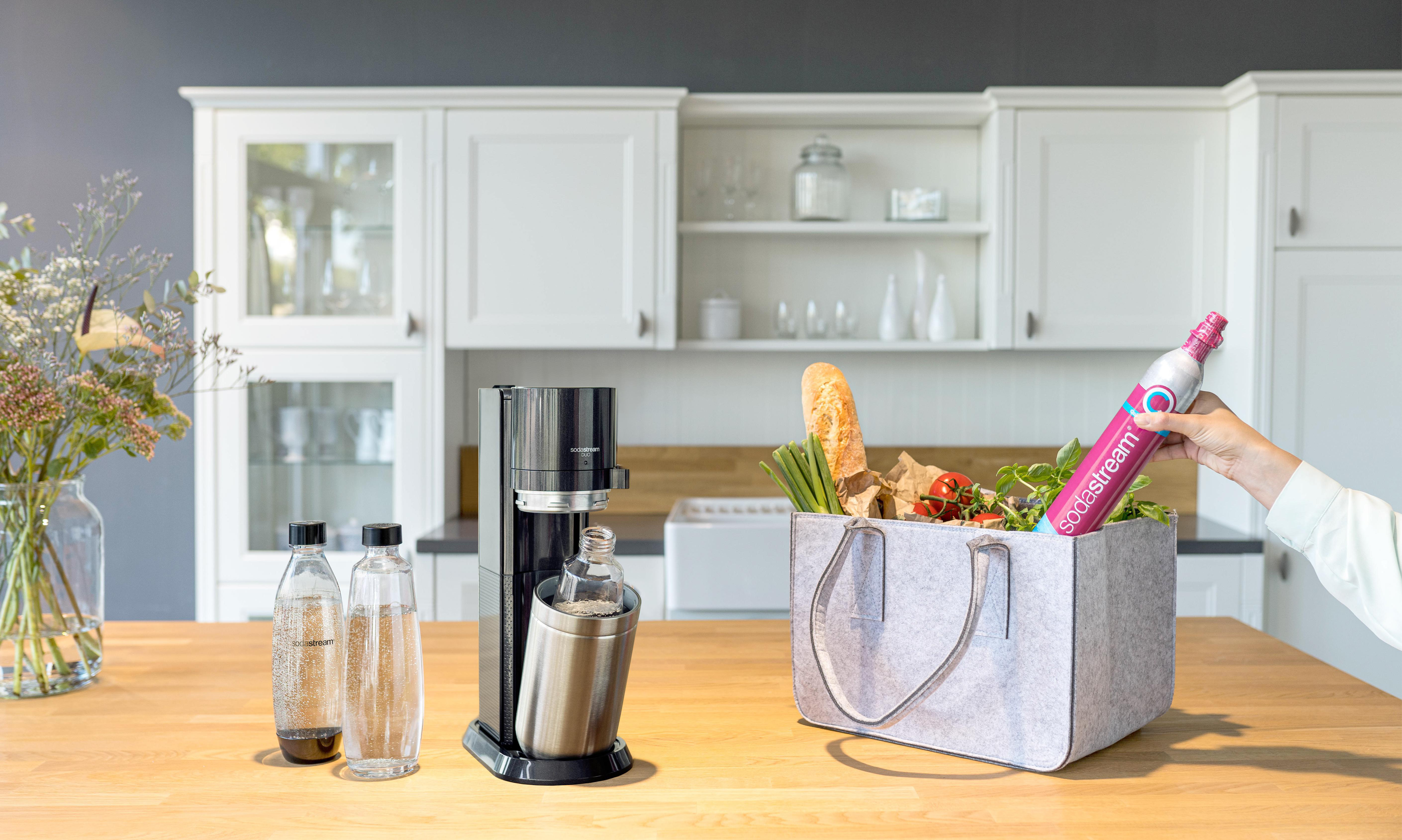 Küchenszene mit Soda-Stream, Flaschen und Lebensmitteln, Person hält Soda-Stream-Kartusche.