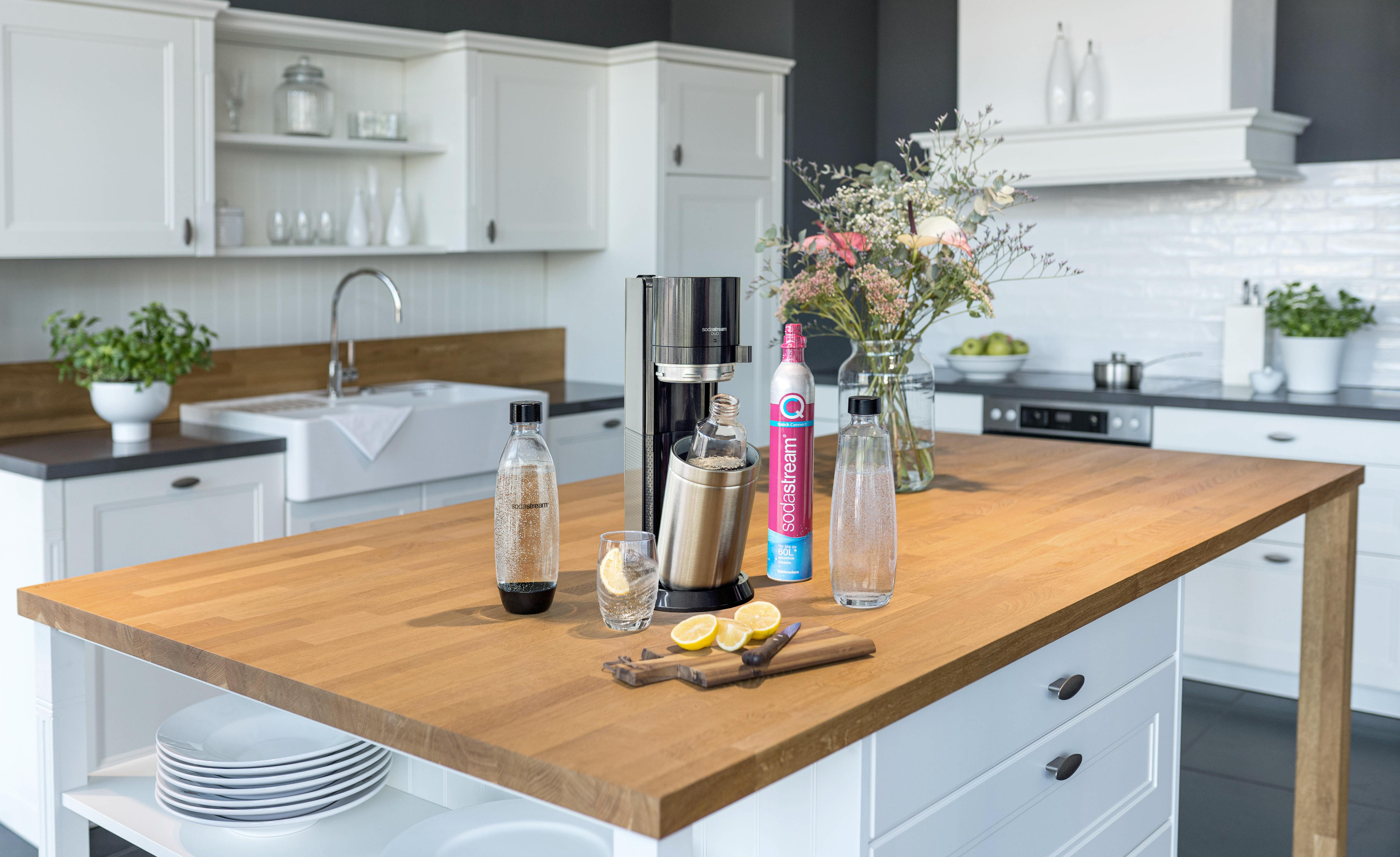 Küchenszene mit Sodamaschine, Flaschen, Zitronen und Blumen auf einer Holzinsel.